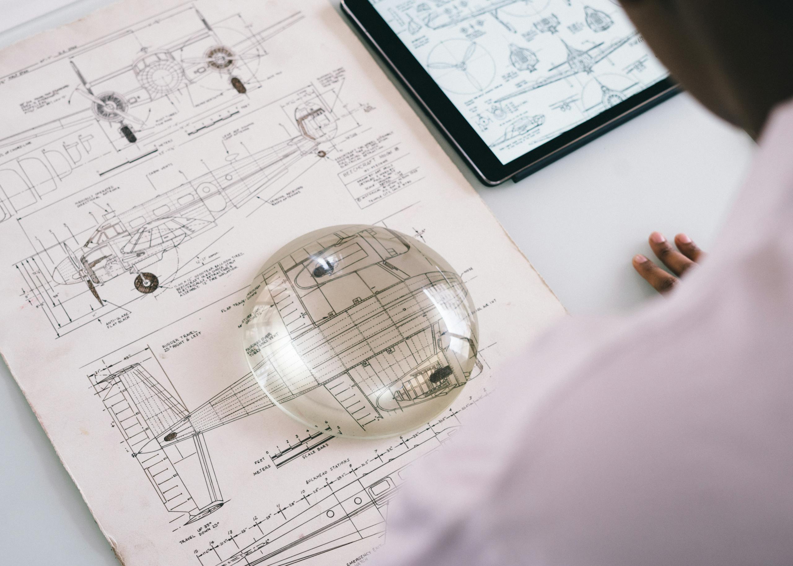 Crop unrecognizable mechanic holding magnifying glass on paper with airplane drawing while working on project on tablet in aircraft factory